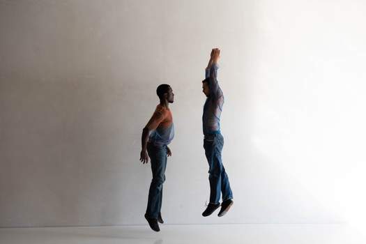 Two dancers jump in parallel lines, one with arms stretched down and the other with arms raised overhead.