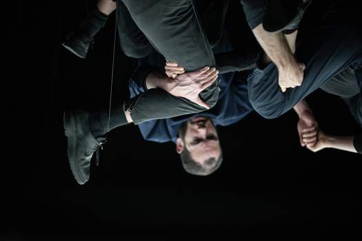 Three performers cling to one another in midair, forming a dense tangle where individual limbs and bodies are barely discernible.
