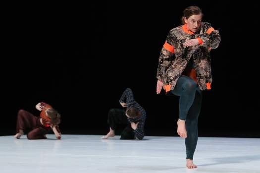 Young dancer stepping as if over an invisible obstacle, focused gaze forward, with two dancers in the background leaning sideways in a crouched pose.