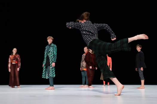 A young dancer caught mid-movement in an energetic, off-balance movement as several other performers look on.