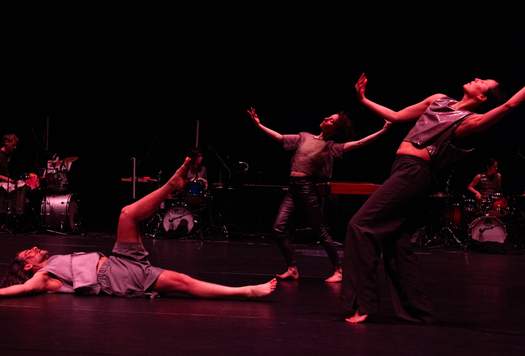 Two dancers mirror each other with arms curved upward in open arcs, while a third dancer lies on the floor with one leg lifted. In the background, three drummers perform among a spread of musical instruments.