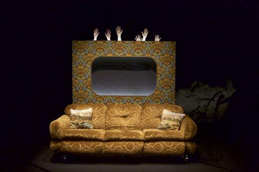 An old coach sits in front of a fabricated backdrop decorated with wallpaper like a grandmother’s house, while six hands carefully emerge from behind the wall.