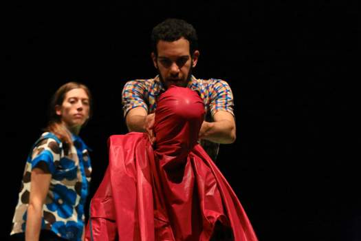 A performer drapes a sheet of plastic over another’s face in a staged, waterboarding-like gesture, while another watches.