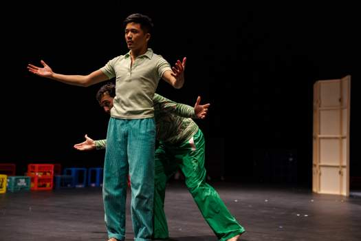 Two performers stand close together as one peeks playfully from behind the other, both extending their arms outward in a warm, welcoming gesture.