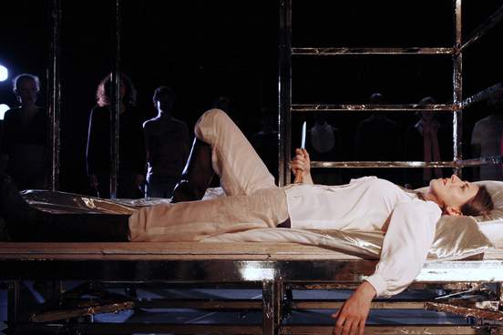 A woman wearing boots lies on a bed-like metal frame, holding a knife upright against her stomach.