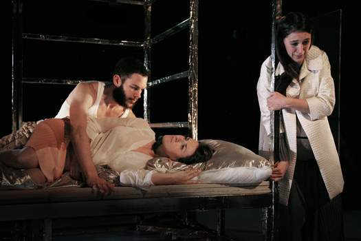 A man leans over a woman lying on a bed-like metal frame, while another woman stands beside them, leaning against the structure with her head bowed as if weeping.