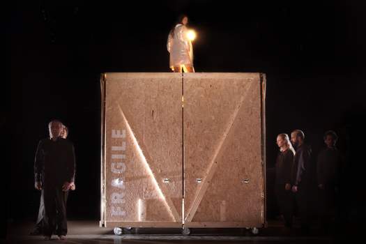 A woman stands atop a large wooden crate marked "FRAGILE", surrounded by several choir members gathered around it.