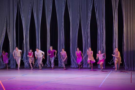 A large group of dancers races from the back to the front of the stage, moving through a curtain of ropes with eager enthusiasm.