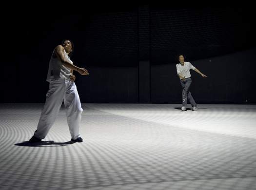 Two dancers move on a bright white stage, the floor patterned with soft, lattice-like shadows created by the lighting.