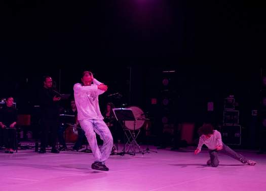 Two dancers in the foreground with instrumentalists and vocalists behind them on a stage lit in pink.
