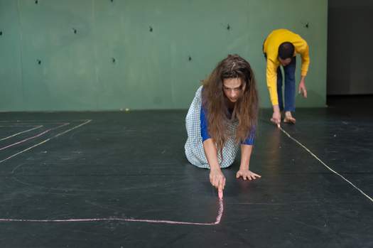 Two actors kneel or bend over as they draw a pattern on the stage floor with colourful chalk.