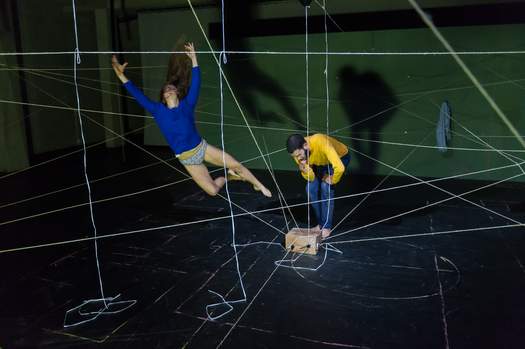 One actor flies through the air mimicking an explosion, while the other crouches in a thoughtful, awkward pose.