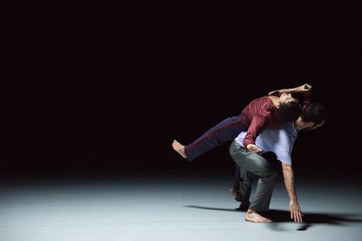 A dancer supports another in a careful balance, backs touching, while the dancer on top maintains a rigid posture.