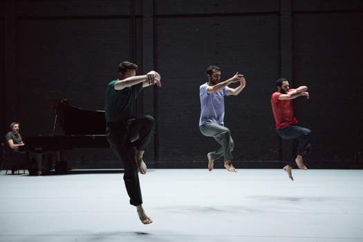 Three dancers are suspended mid-air, about to land from a unison jump, arms lifted to chest height, while a pianist watches from the back corner of the stage.