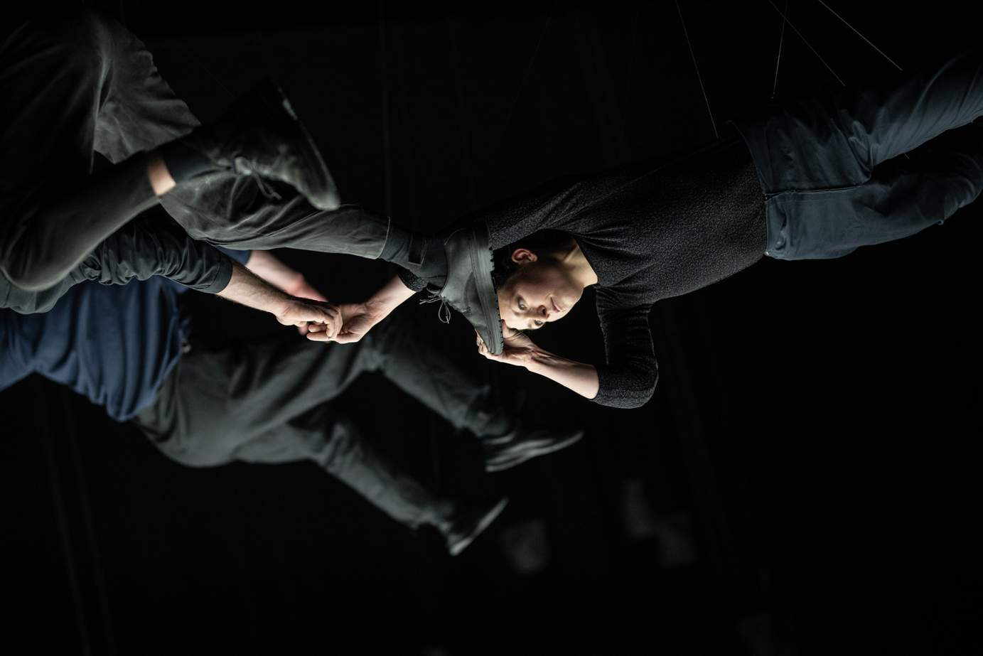 Three performers hang in midair by thin ropes in a dark space lit from below; two of them cling to each other by hands and feet.
