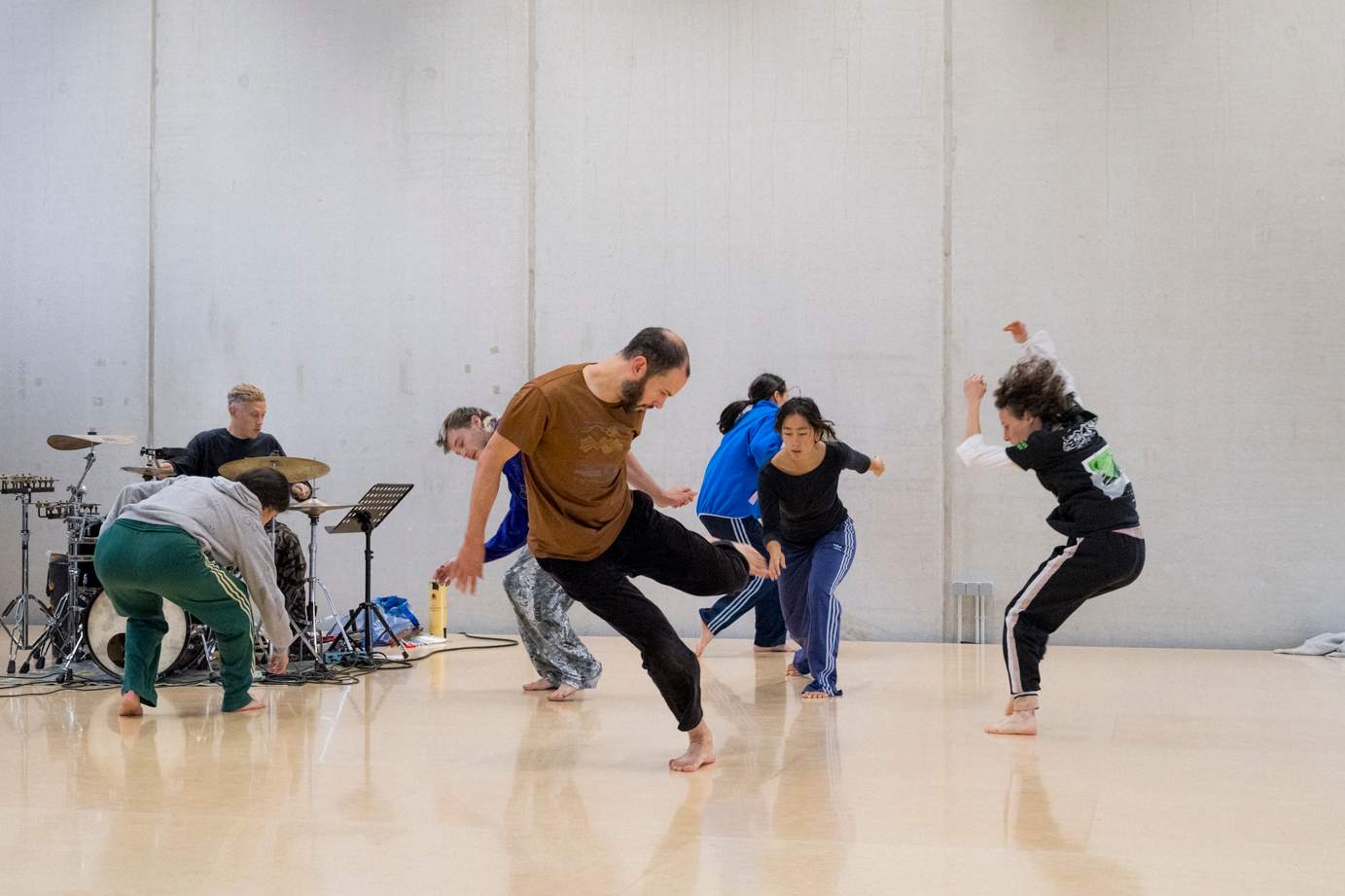 Six dancers practice energetic choreography during a rehearsal as a drummer provides live rhythm.