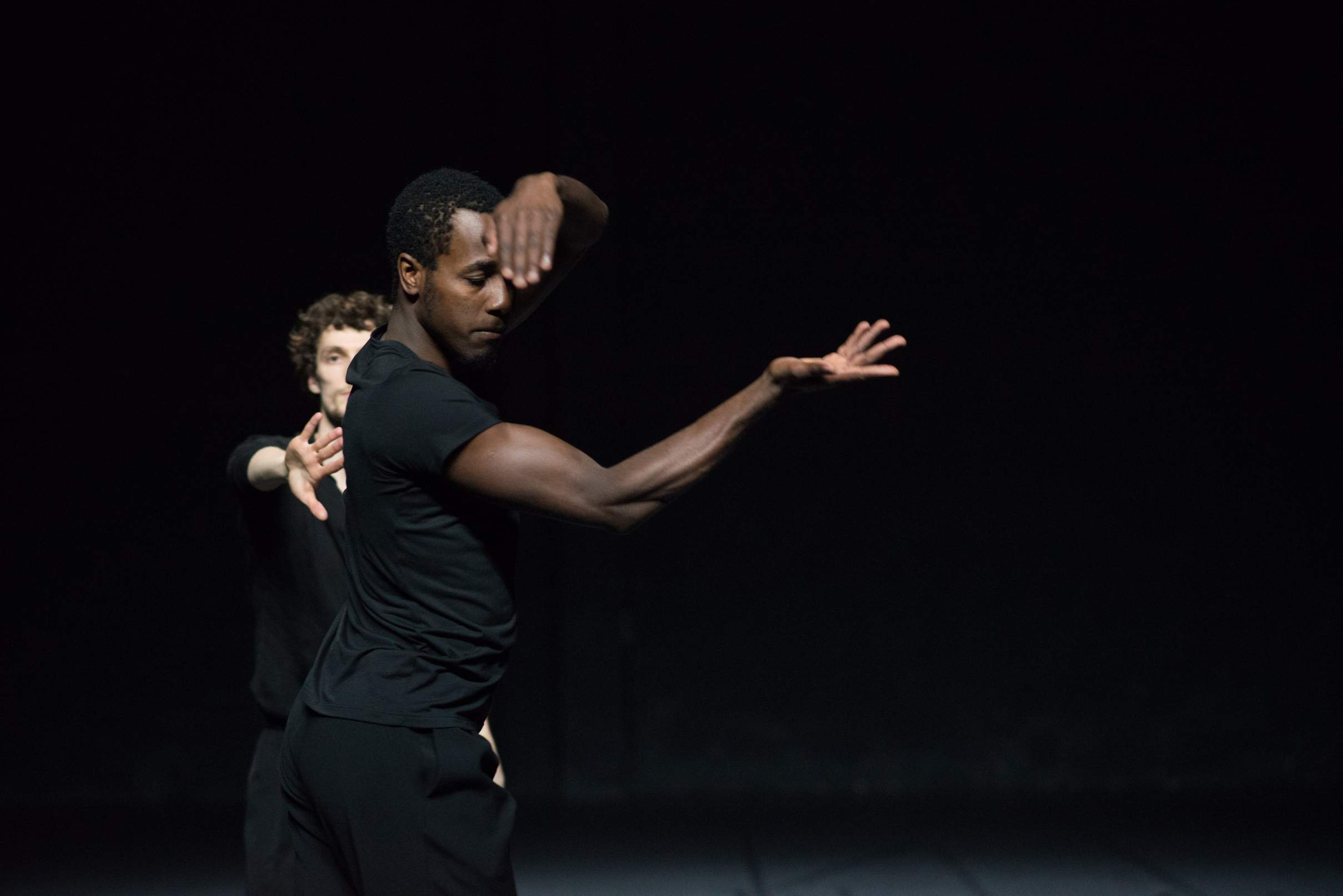 One dancer moves his arms in a circular motion, while another behind him raises a hand forward toward the audience.