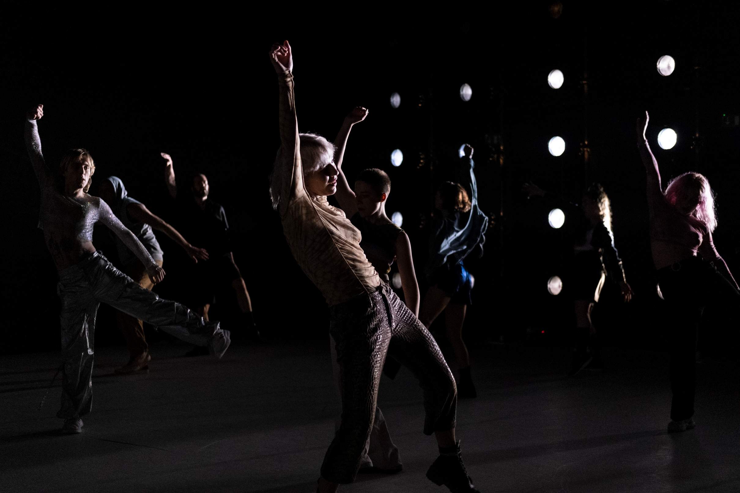 Several dancers perform a synchronized movement in a dimly lit room, lifting one arm into the air and shifting their hips in unison.