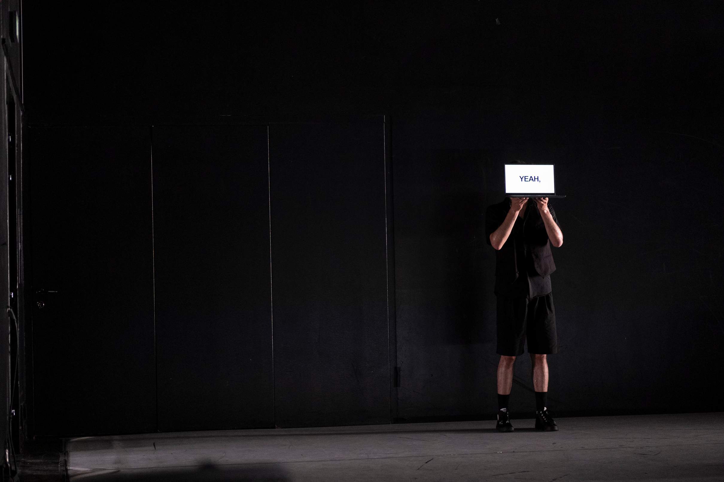A performer stands against the back wall, holding a laptop screen in front of their face that reads “YEAH” in all caps.