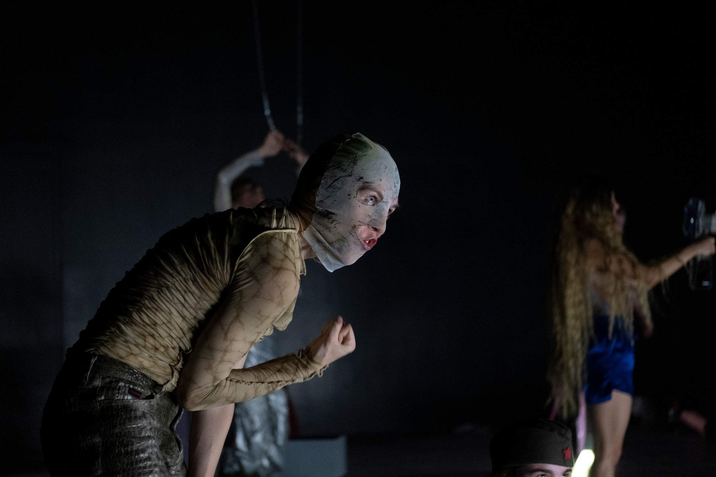 Three performers carry out different actions across the stage. The performer in the foreground faces the audience, her face concealed by a balaclava.