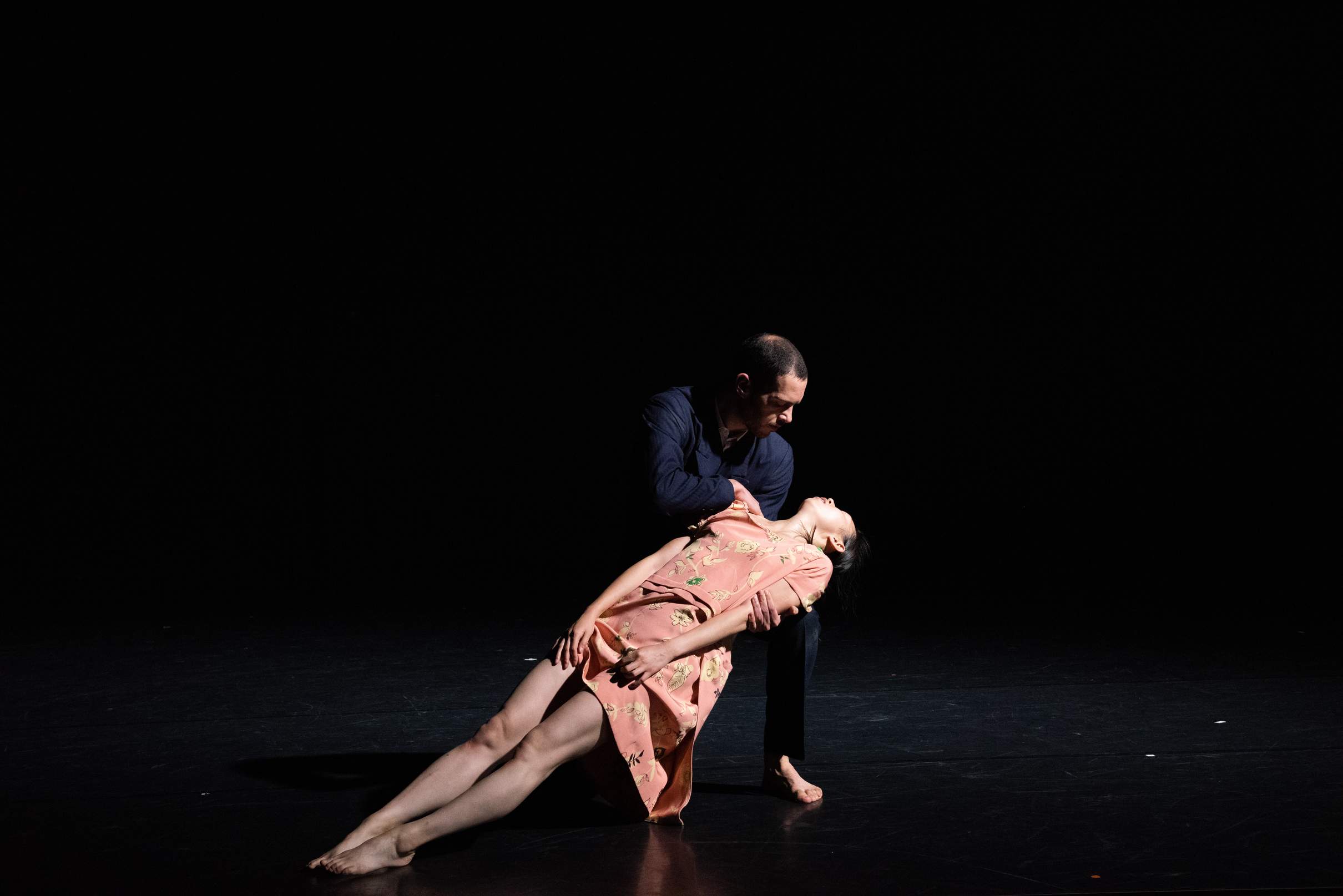 A male performer in a suit carries a female performer in a dress on his knee, her body fully outstretched like a plank.