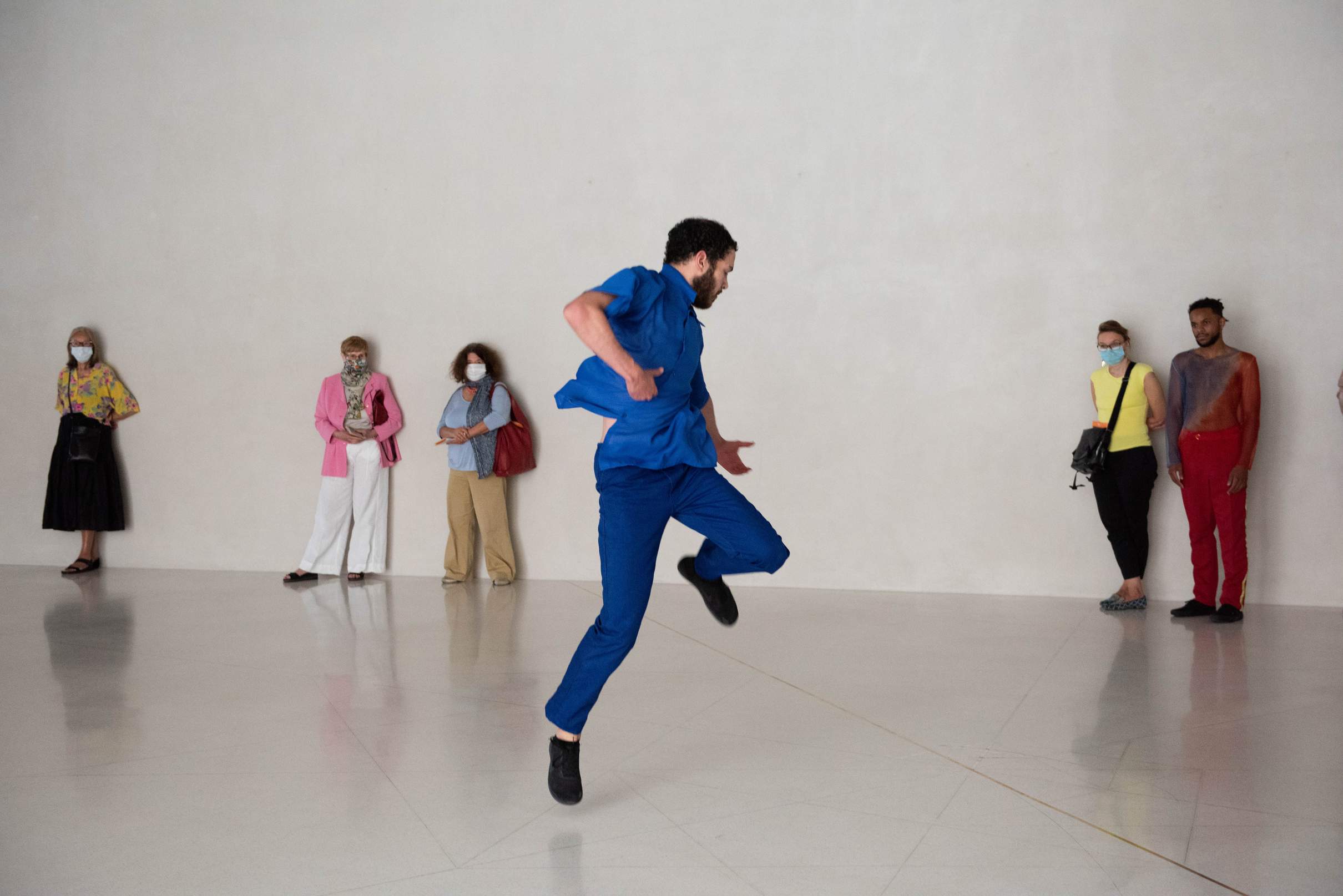 A dancer leaps into the air, observed by another dancer and a group of masked museum visitors standing along the wall.