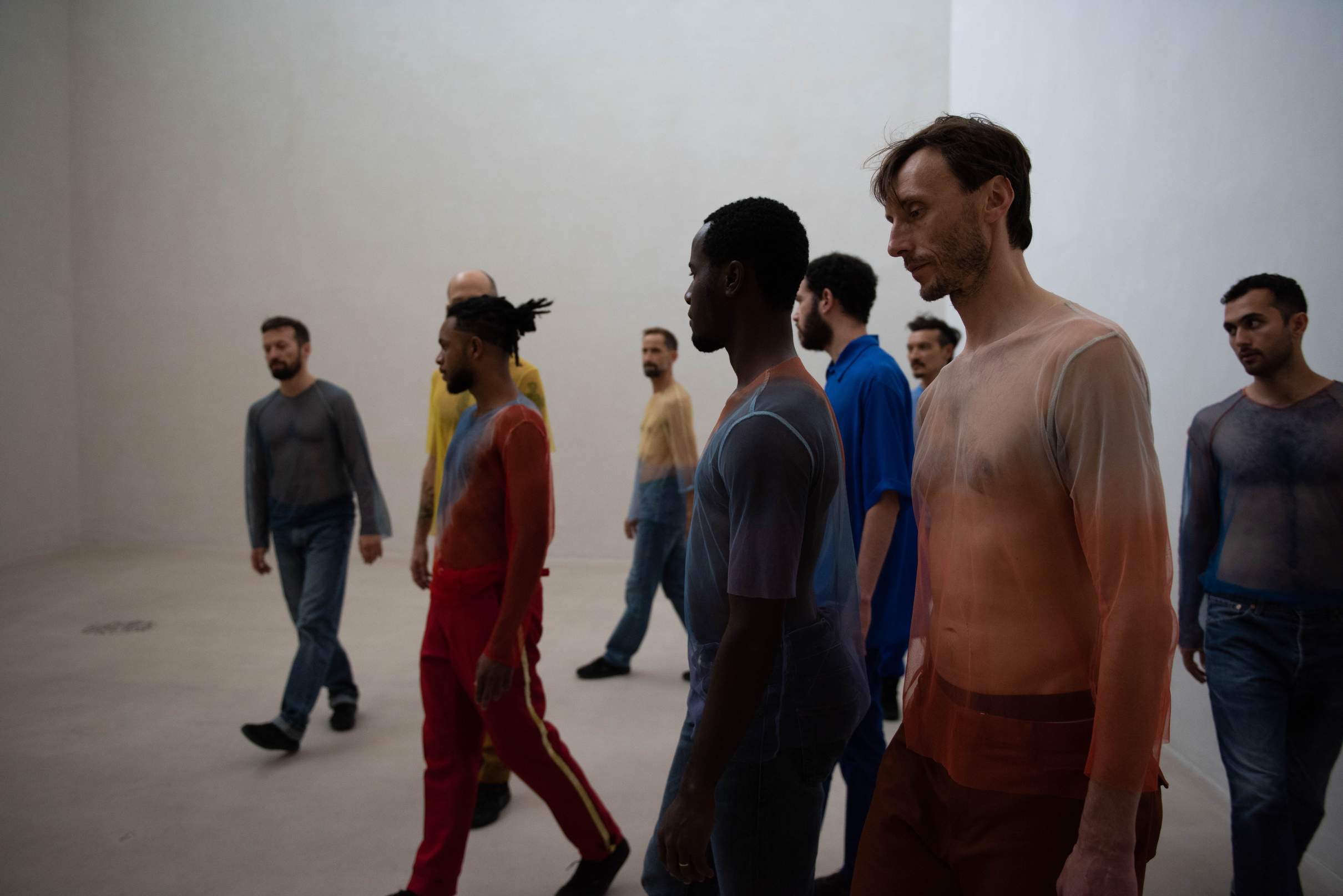 A large group of male performers weave among one another while walking through a spacious, statuesque museum hall.