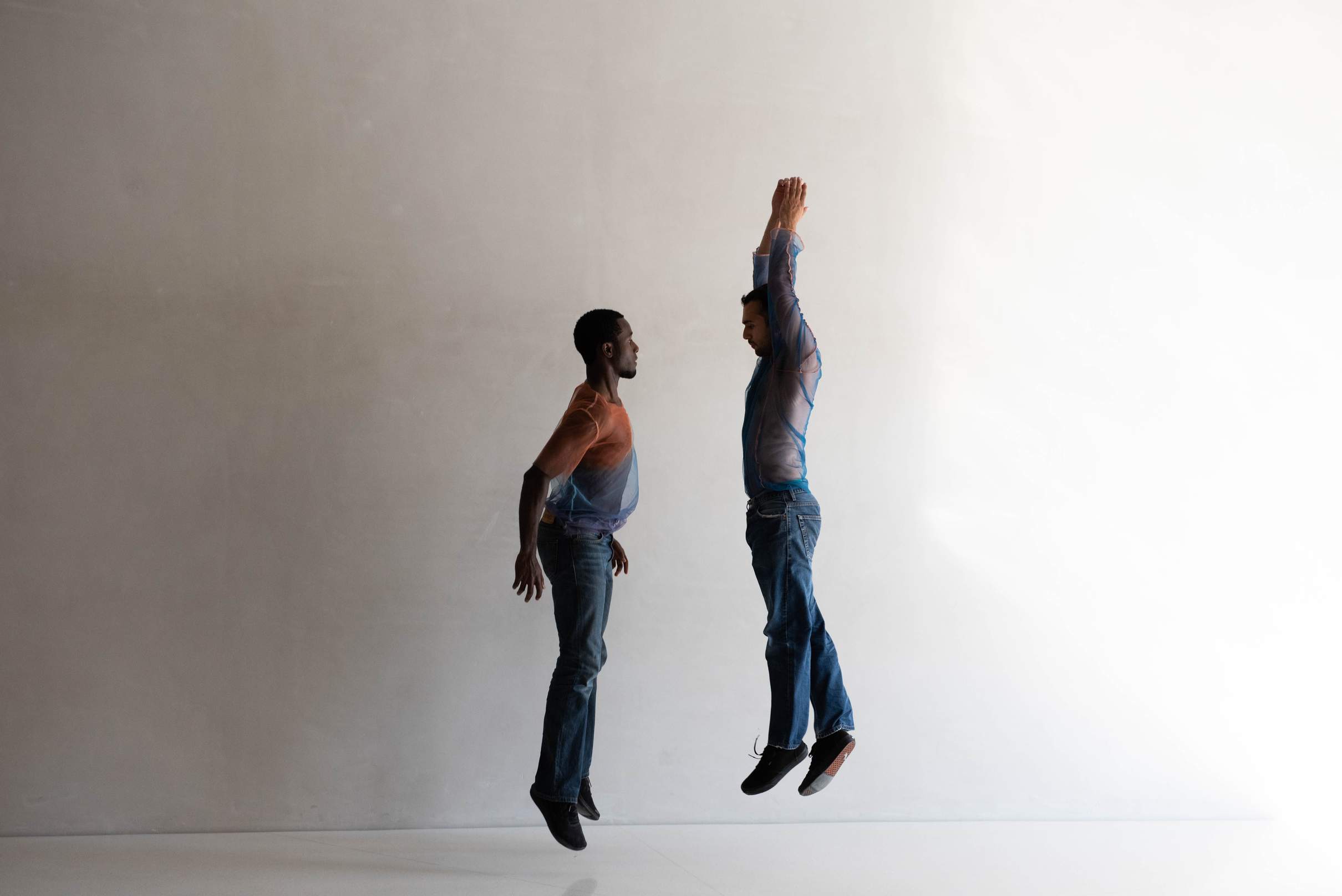 Two dancers jump in parallel lines, one with arms stretched down and the other with arms raised overhead.