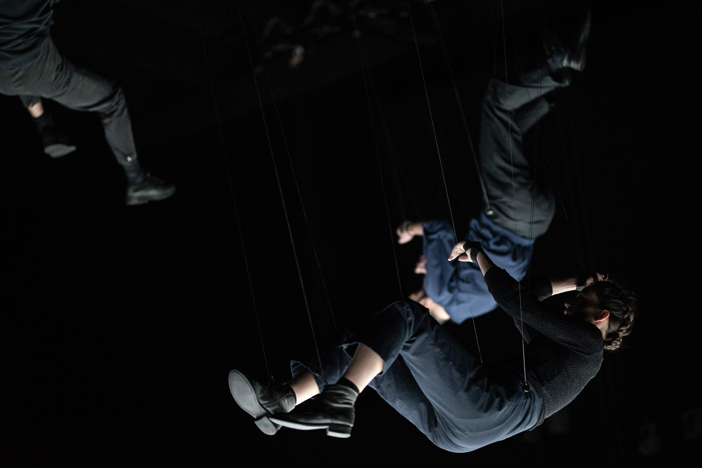 Three performers hang in midair on thin ropes, each in a different orientation and posture.