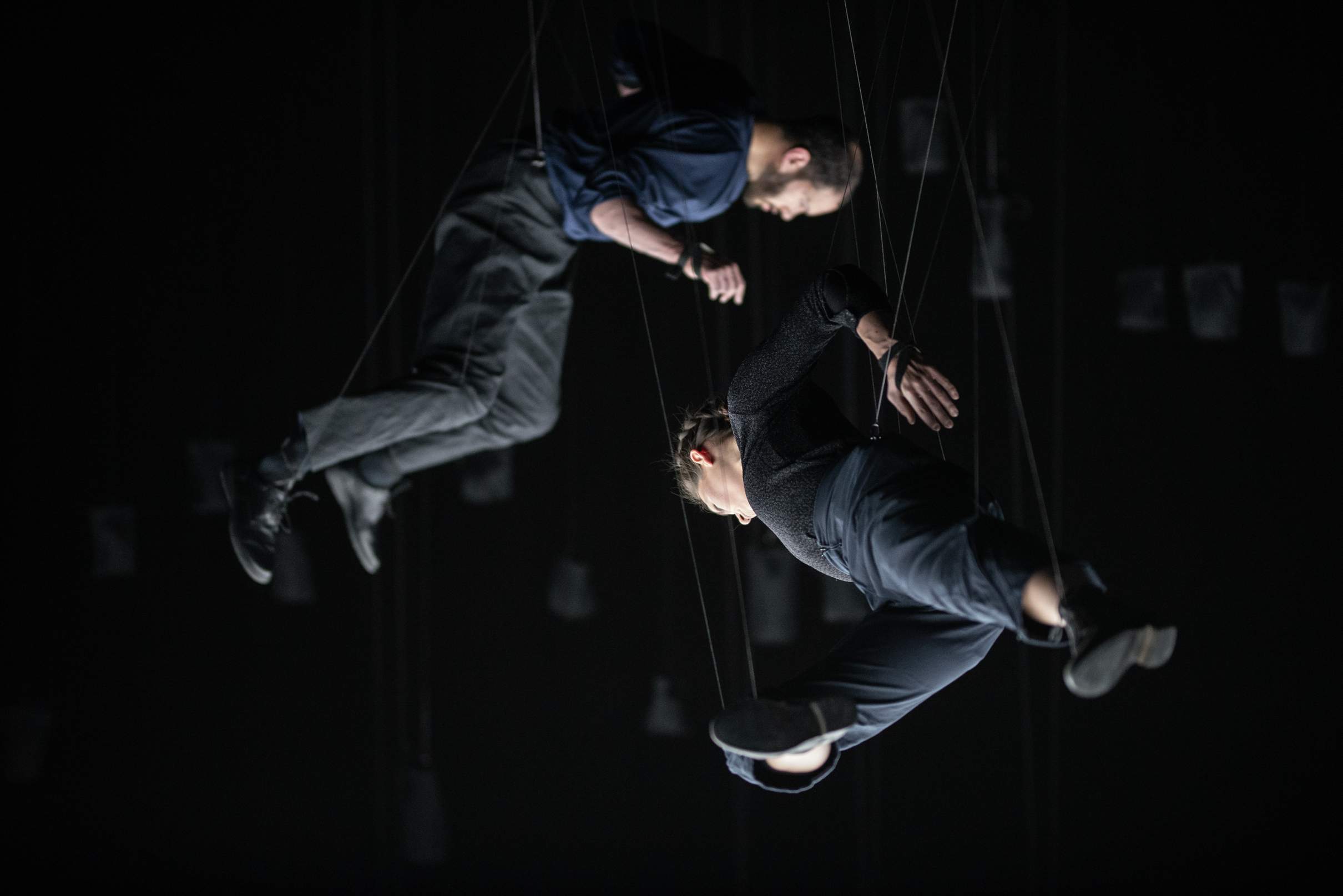 Two performers hang in midair on thin ropes, facing downward as if flying above the floor, with counterweight bags visible behind them.
