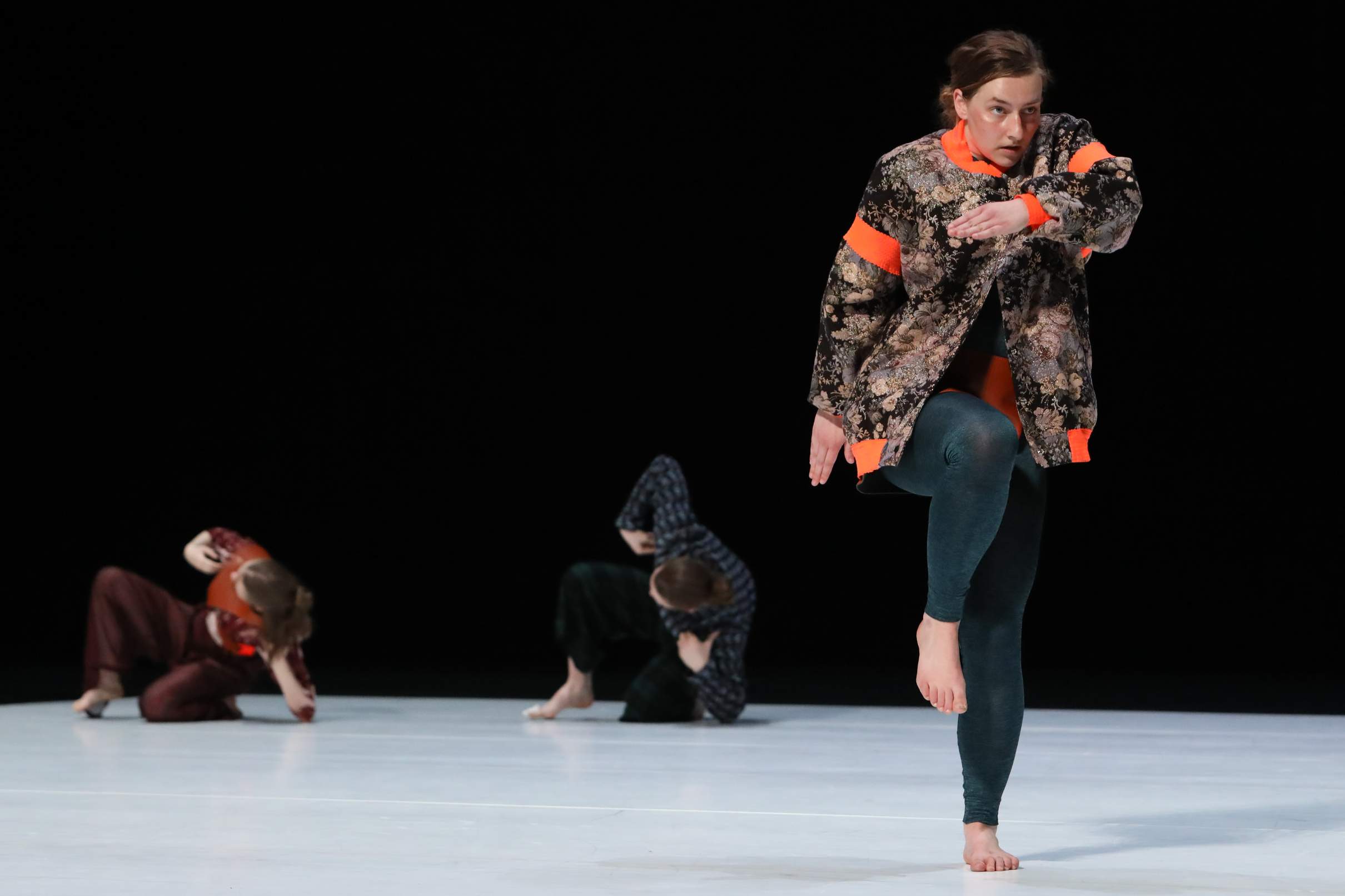 Young dancer stepping as if over an invisible obstacle, focused gaze forward, with two dancers in the background leaning sideways in a crouched pose.