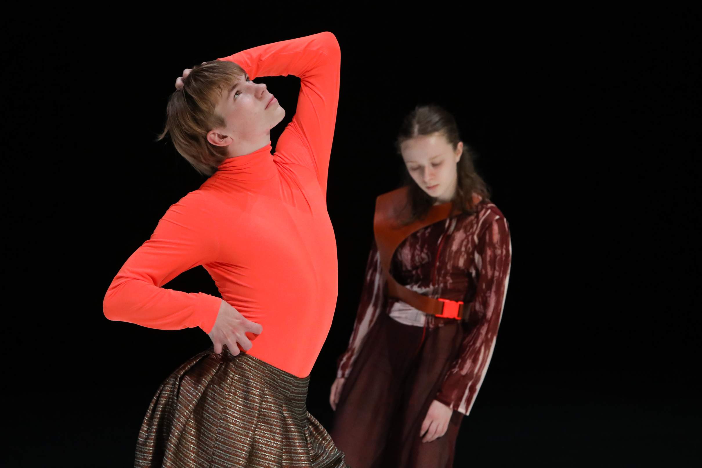 A young dancer in a fluorescent orange costume poses prominently, while a second dancer in the background appears more withdrawn.