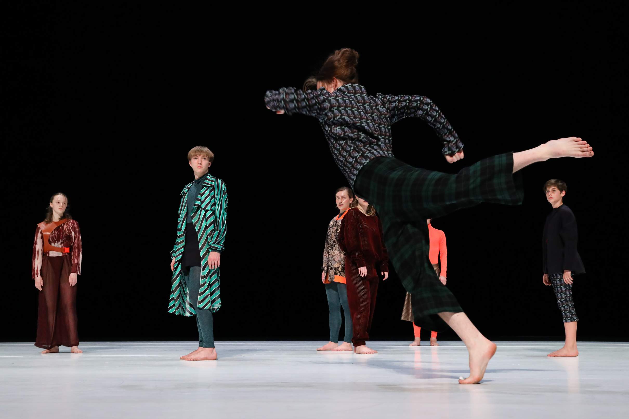 A young dancer caught mid-movement in an energetic, off-balance movement as several other performers look on.
