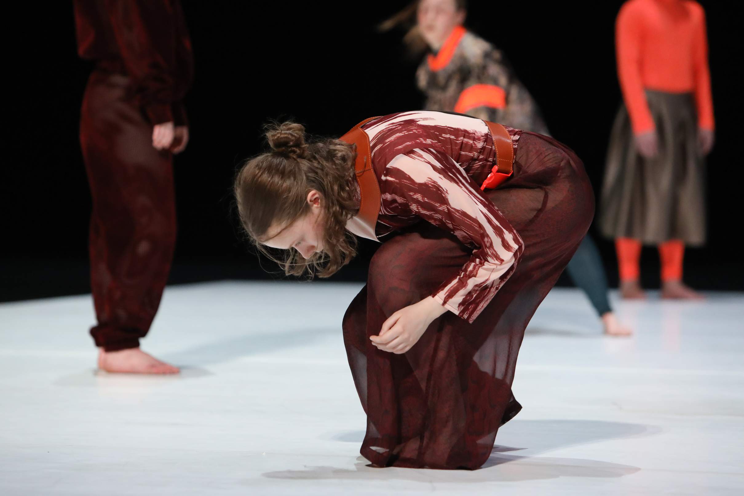 A young dancer bends forward in a half-crouch, facing the white dance floor, with three other dancers in the background.