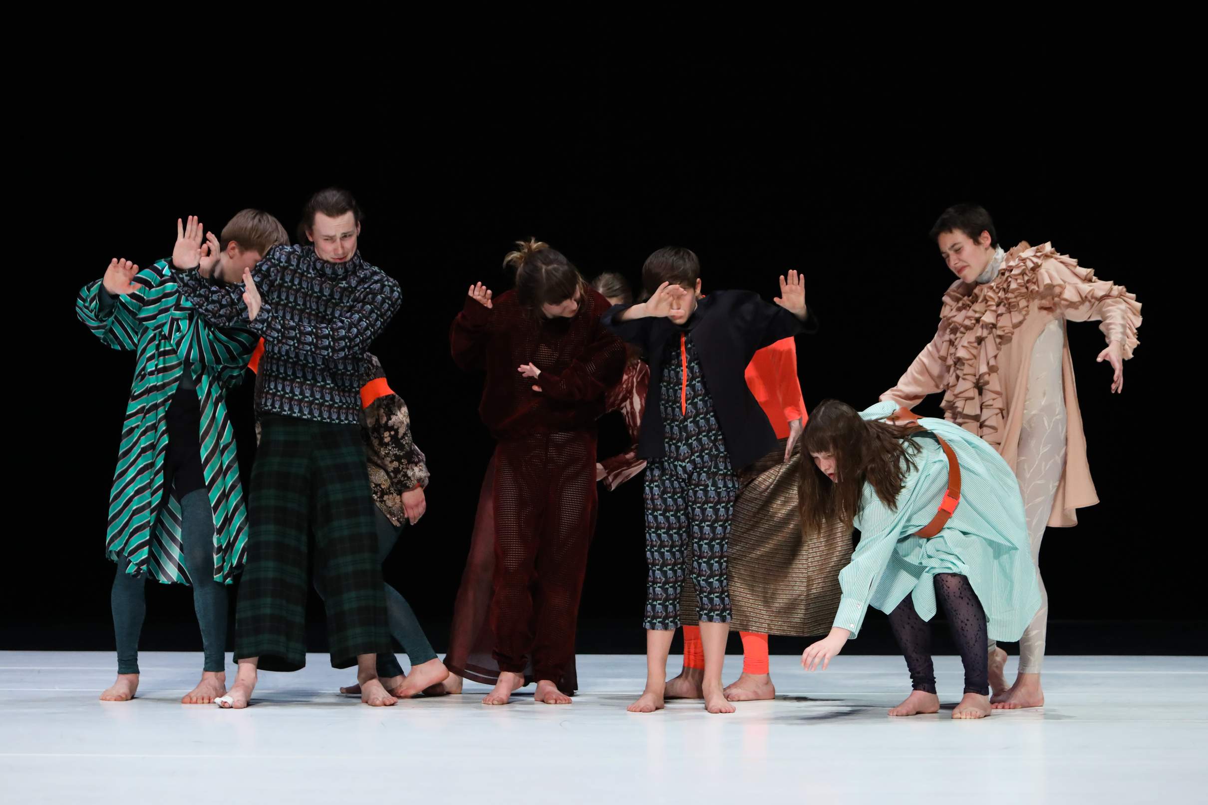 Nine young performers cluster tightly in a group, their bodies and gestures conveying aversion, discomfort, and distress.
