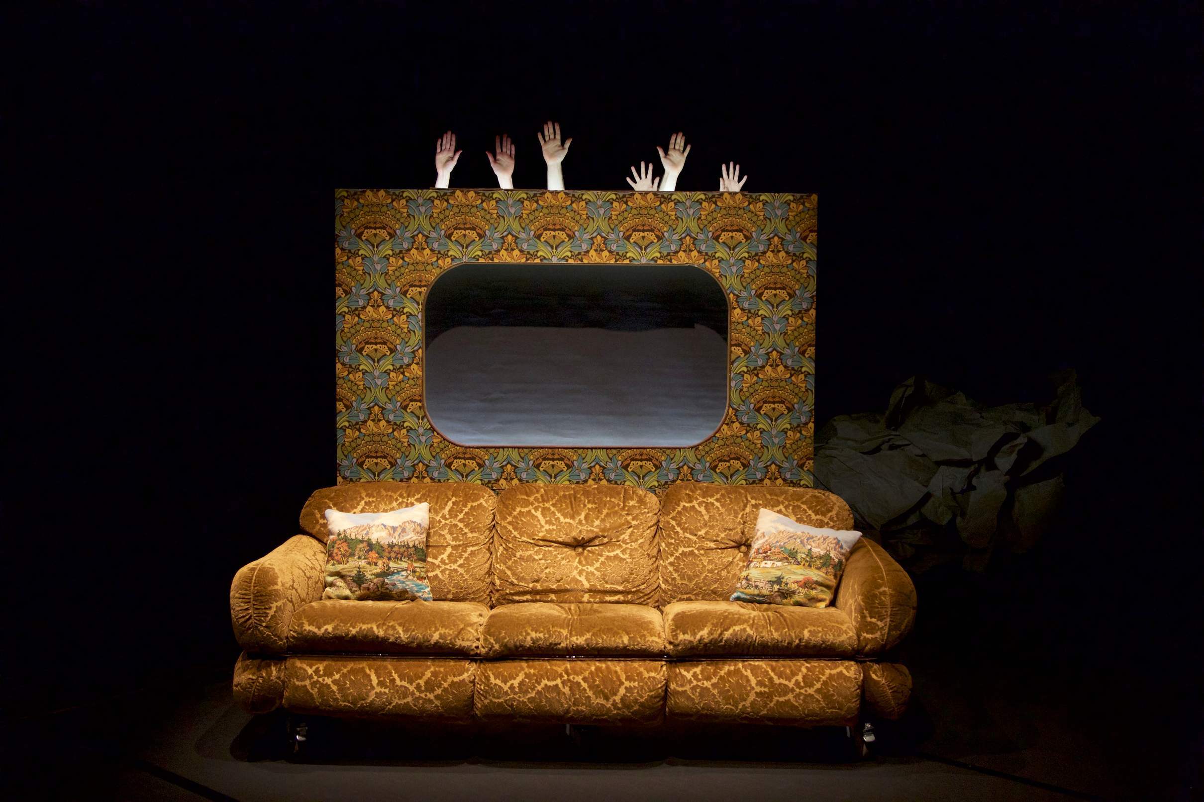 An old coach sits in front of a fabricated backdrop decorated with wallpaper like a grandmother’s house, while six hands carefully emerge from behind the wall.