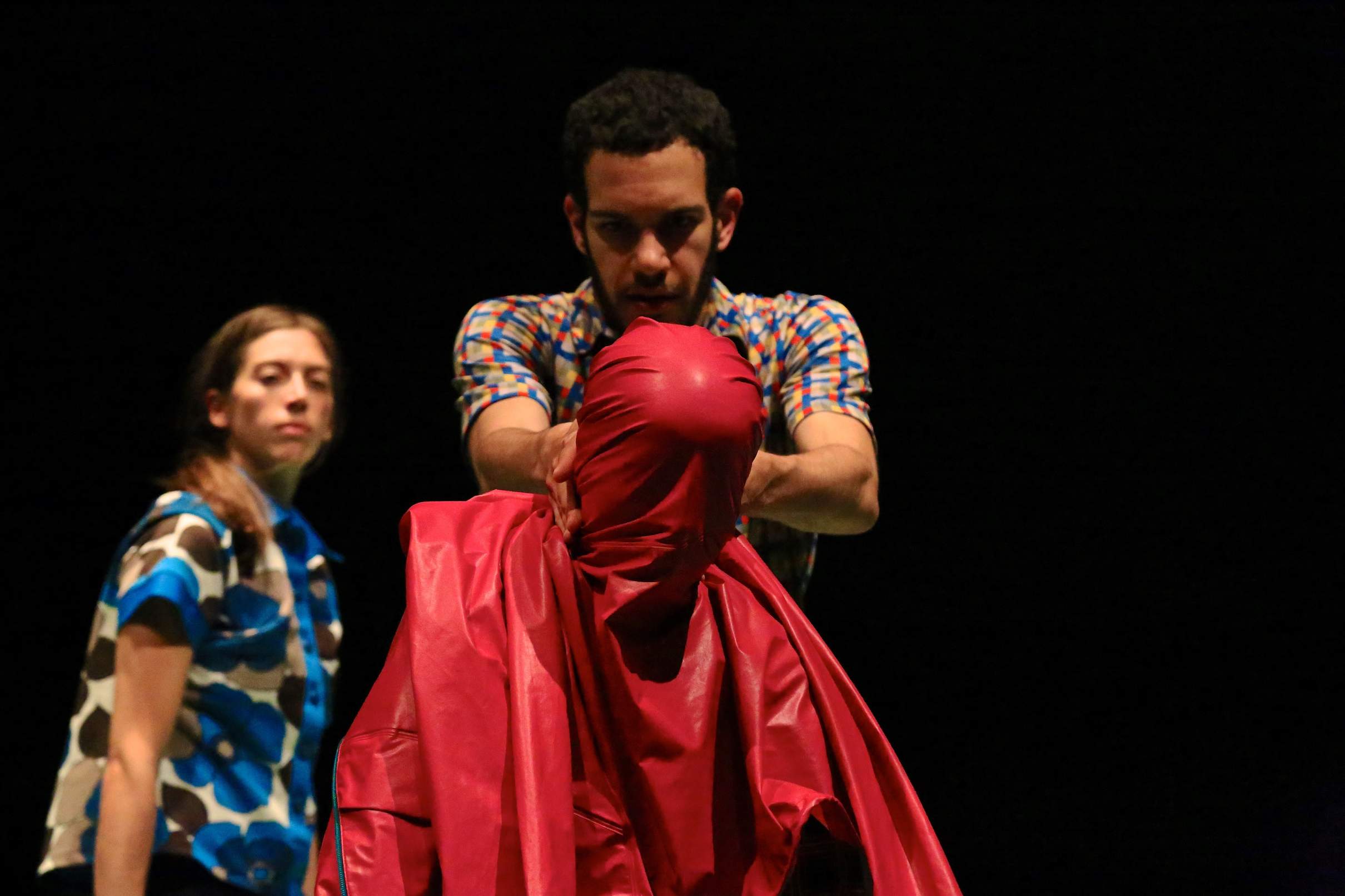 A performer drapes a sheet of plastic over another’s face in a staged, waterboarding-like gesture, while another watches.