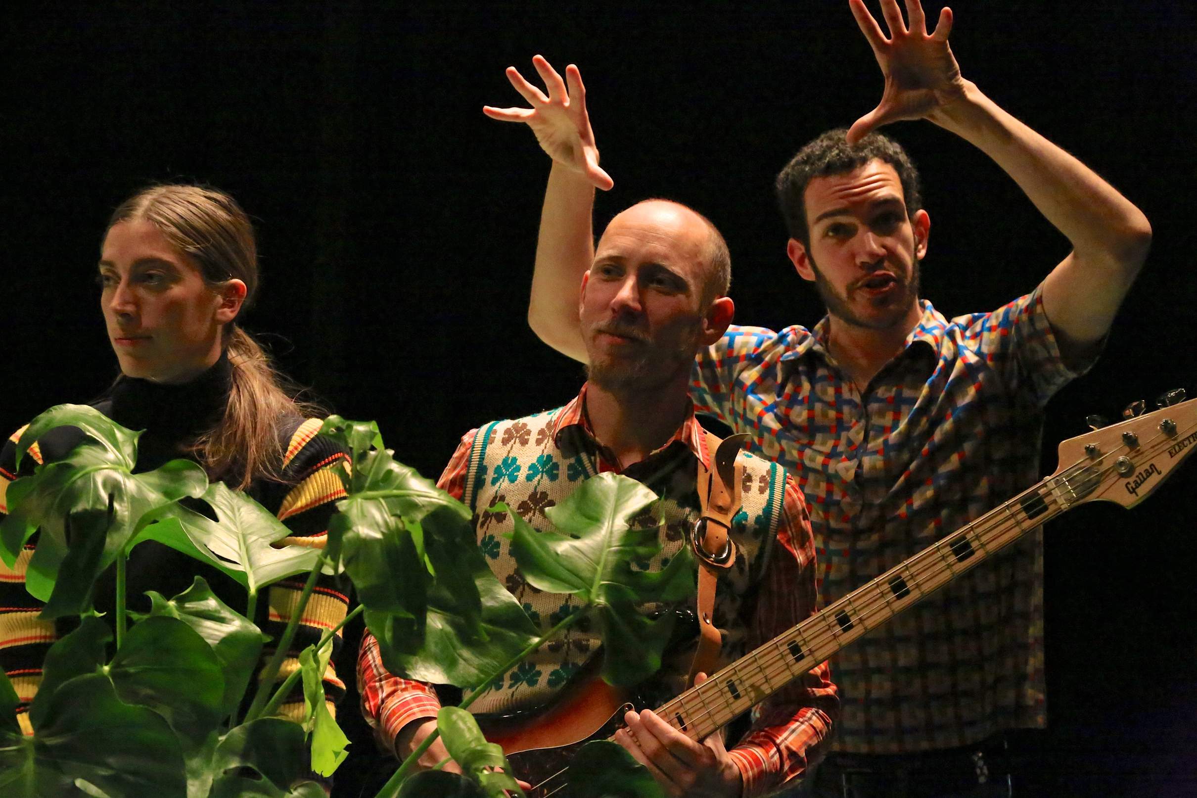 Three performers stand close together: one holding a plant, one holding a guitar, and the third with hands raised, addressing the audience.
