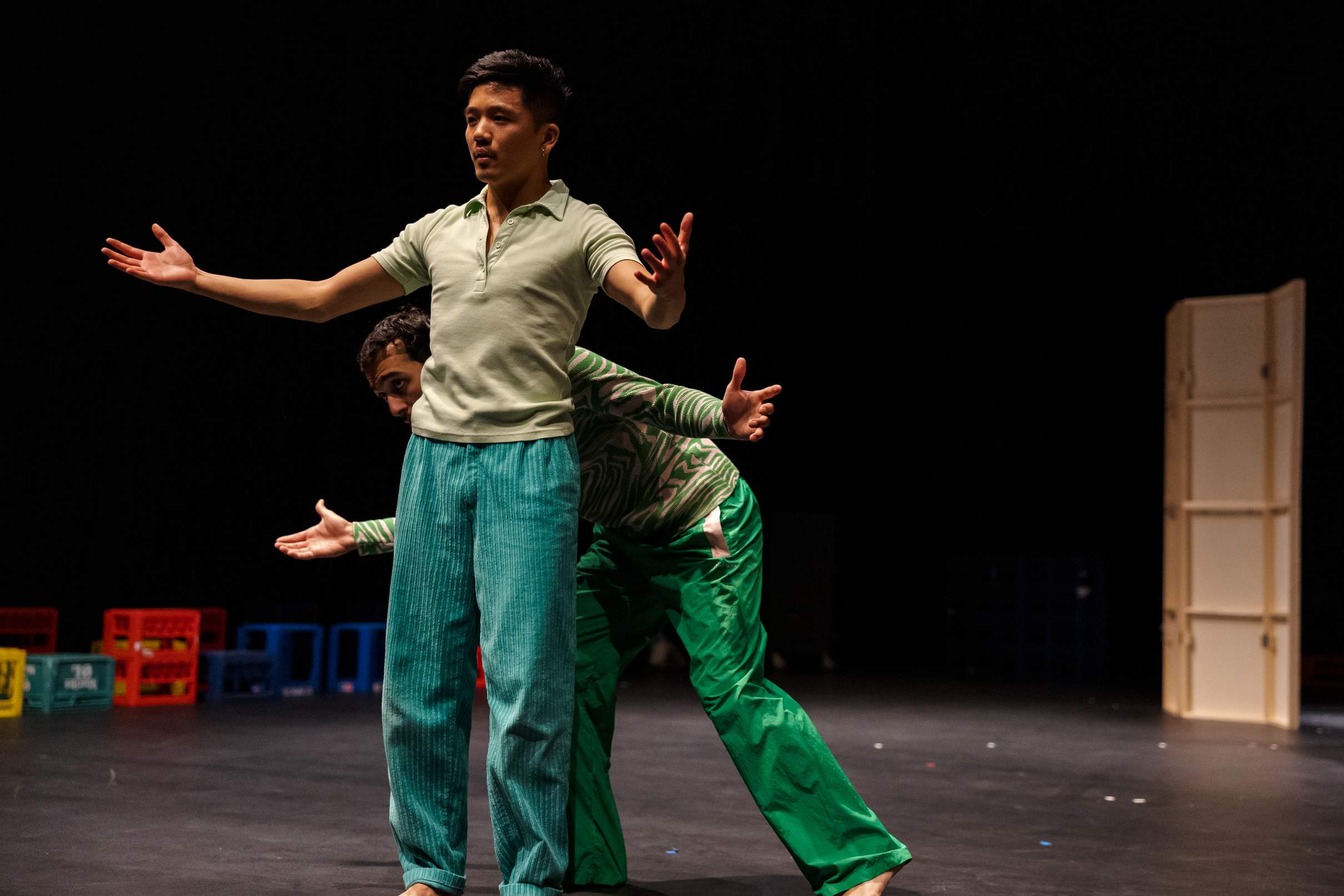 Two performers stand close together as one peeks playfully from behind the other, both extending their arms outward in a warm, welcoming gesture.