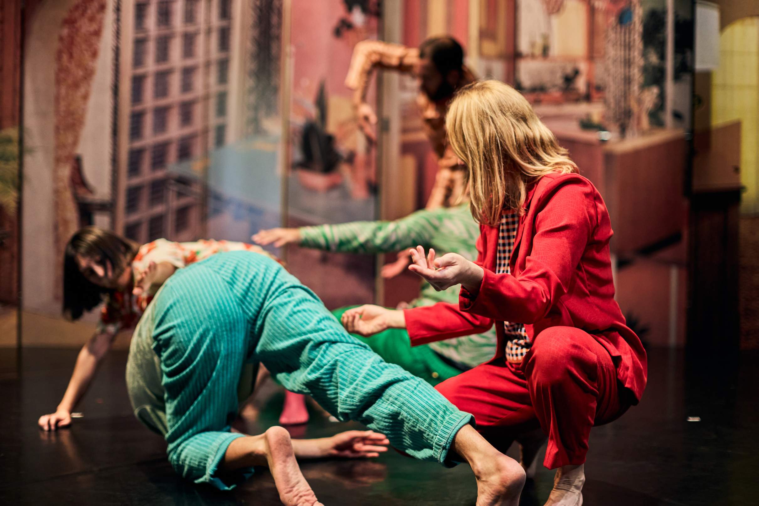 Five performers in colorful outfits twist and turn their bodies to form a living blueprint, set against a backdrop of a wall decorated with retro furniture and patterned wallpaper.