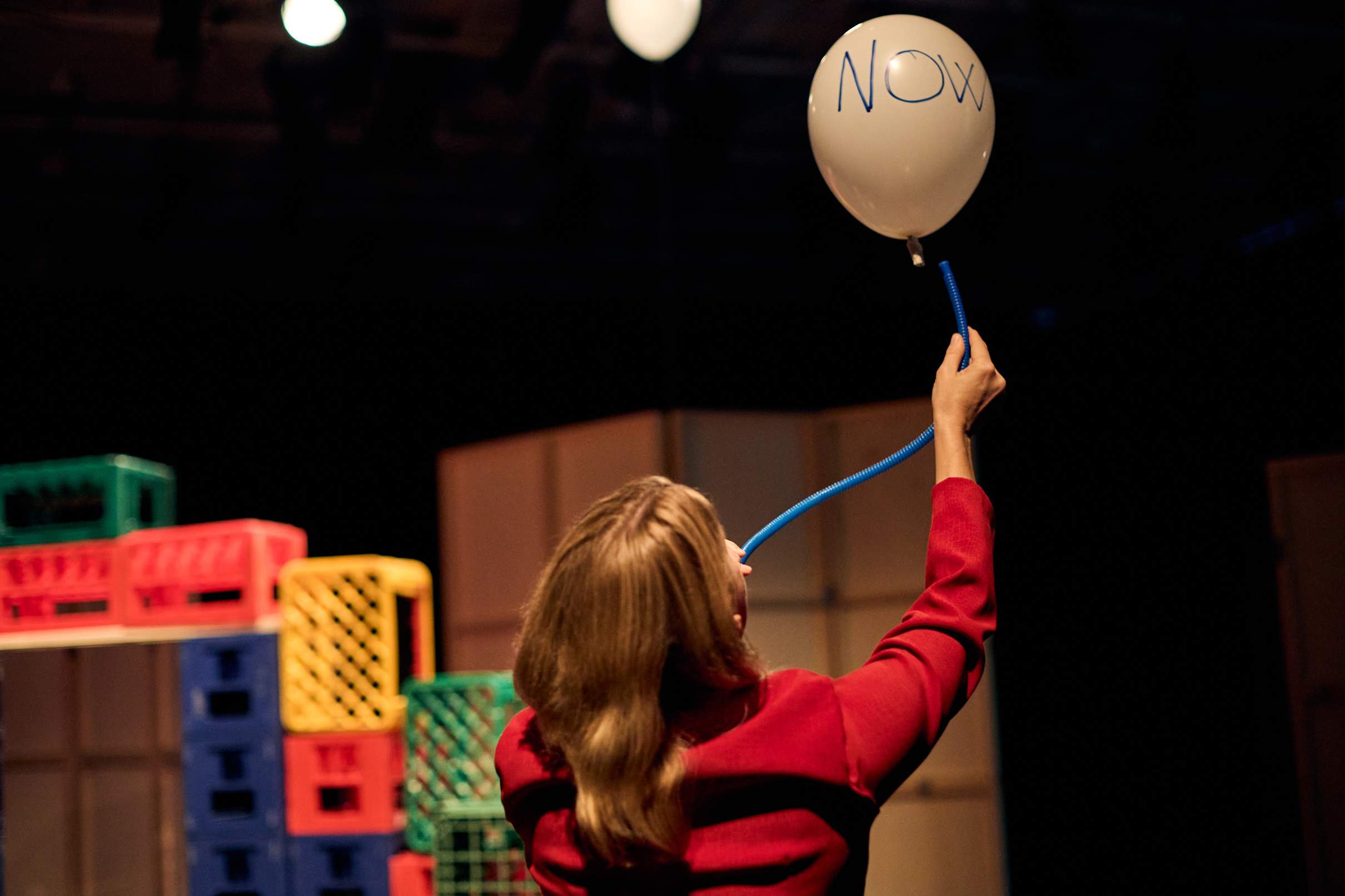 A performer in a red outfit blows air through a blue tube to keep a balloon labeled NOW floating in the air.
