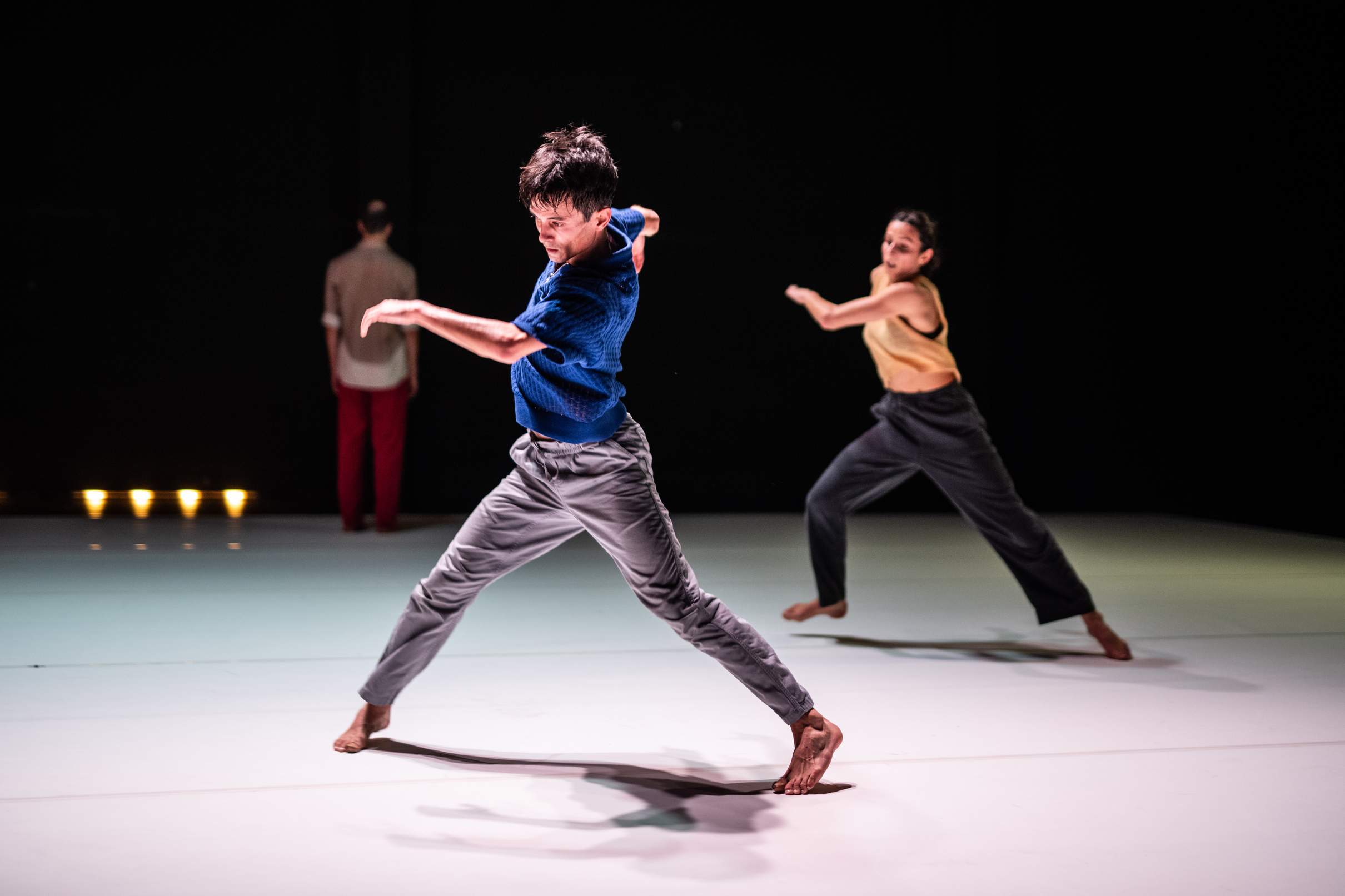 Two sweat-drenched dancers perform a vigorous dance as another dancer stands in the background facing away.