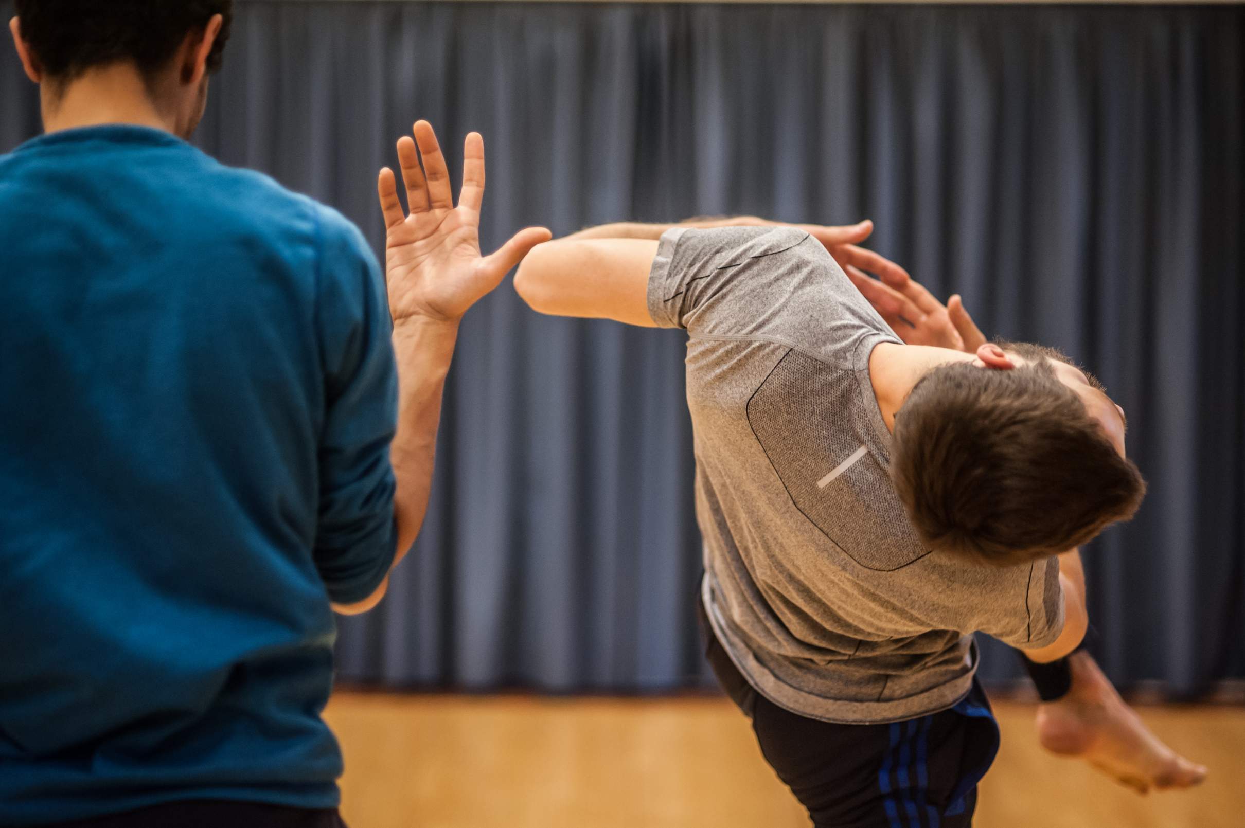 Two dancers, close together and facing back, perform contrasting movements: one stands with a hand raised to shoulder height, the other in a strained, tilted twist.