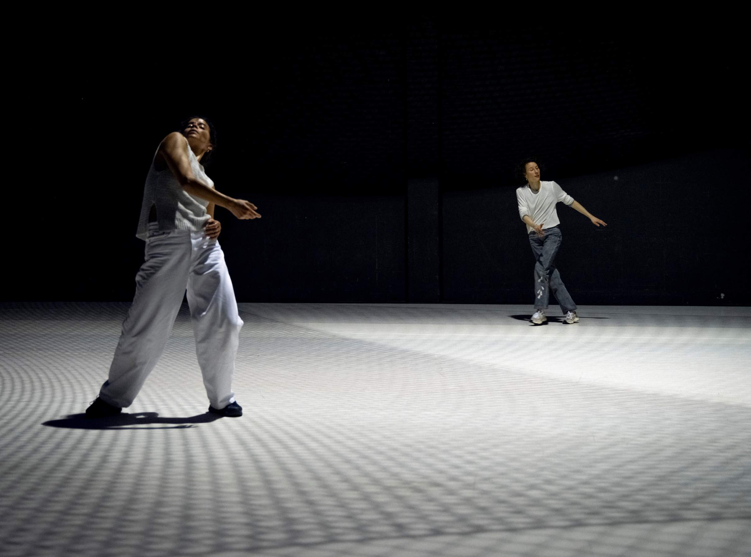 Two dancers move on a bright white stage, the floor patterned with soft, lattice-like shadows created by the lighting.