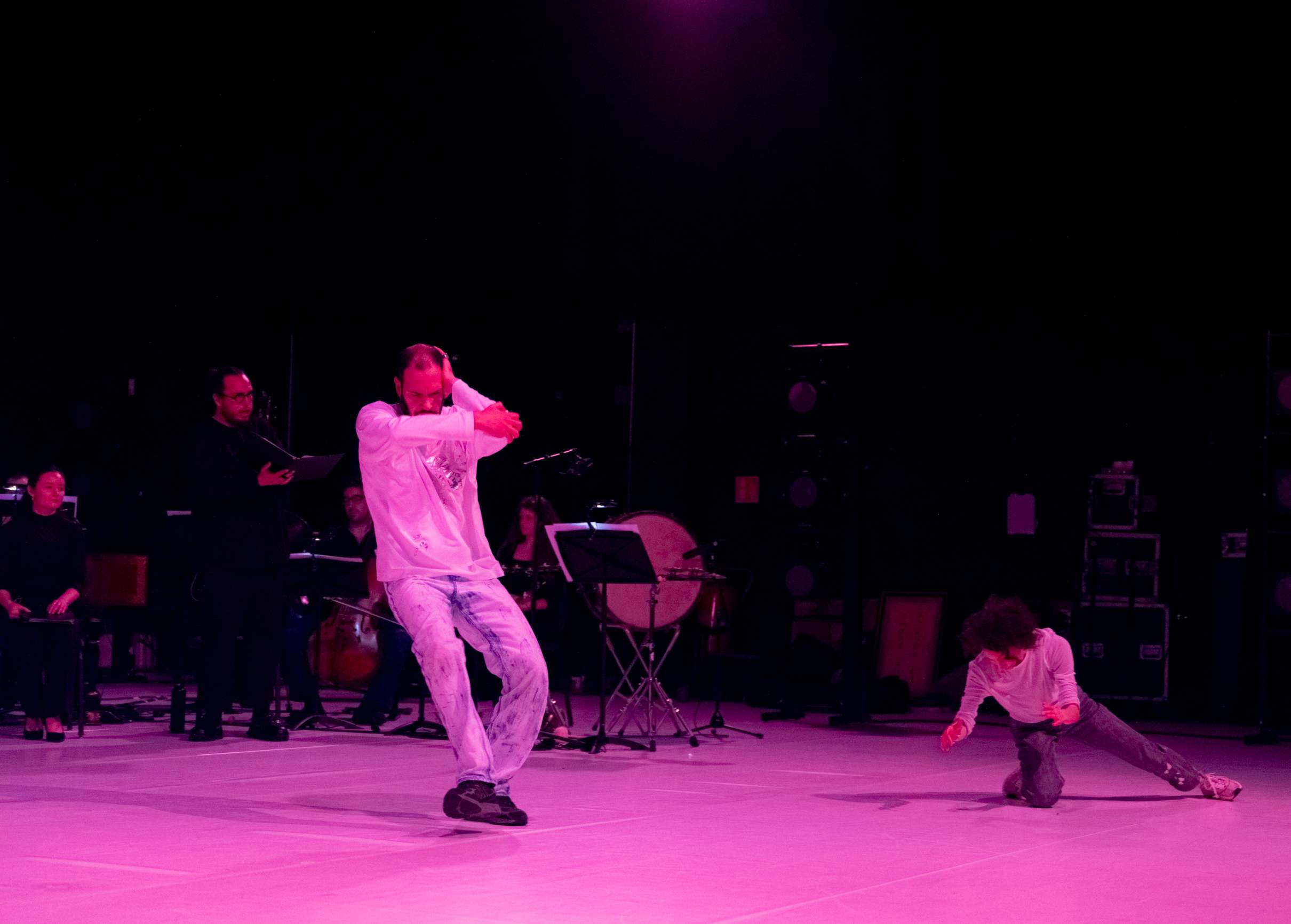 Two dancers in the foreground with instrumentalists and vocalists behind them on a stage lit in pink.