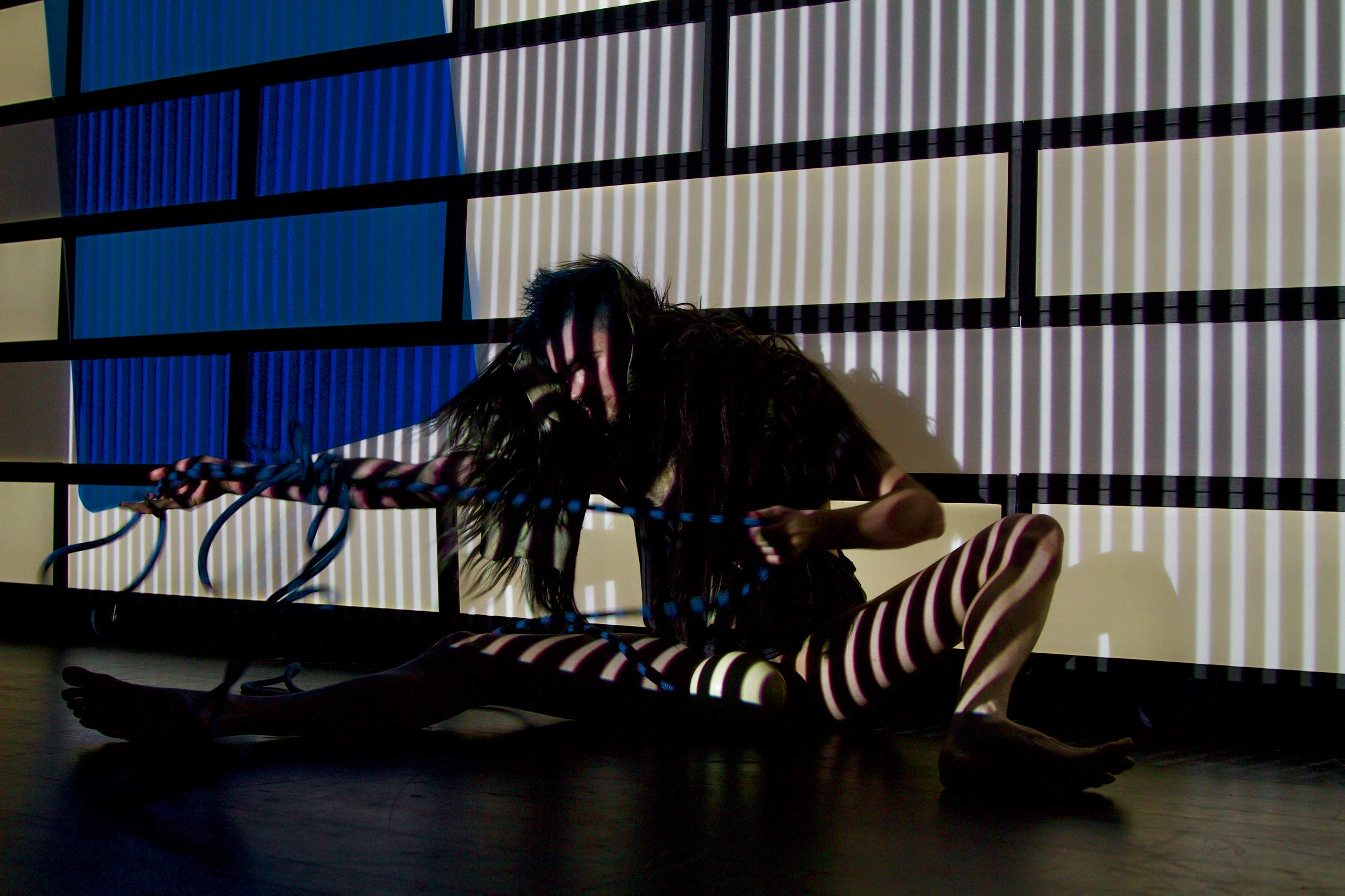 A performer sits against a patterned wall striped with bands of light and shadow, holding a large egg between their legs while struggling with a blue rope.