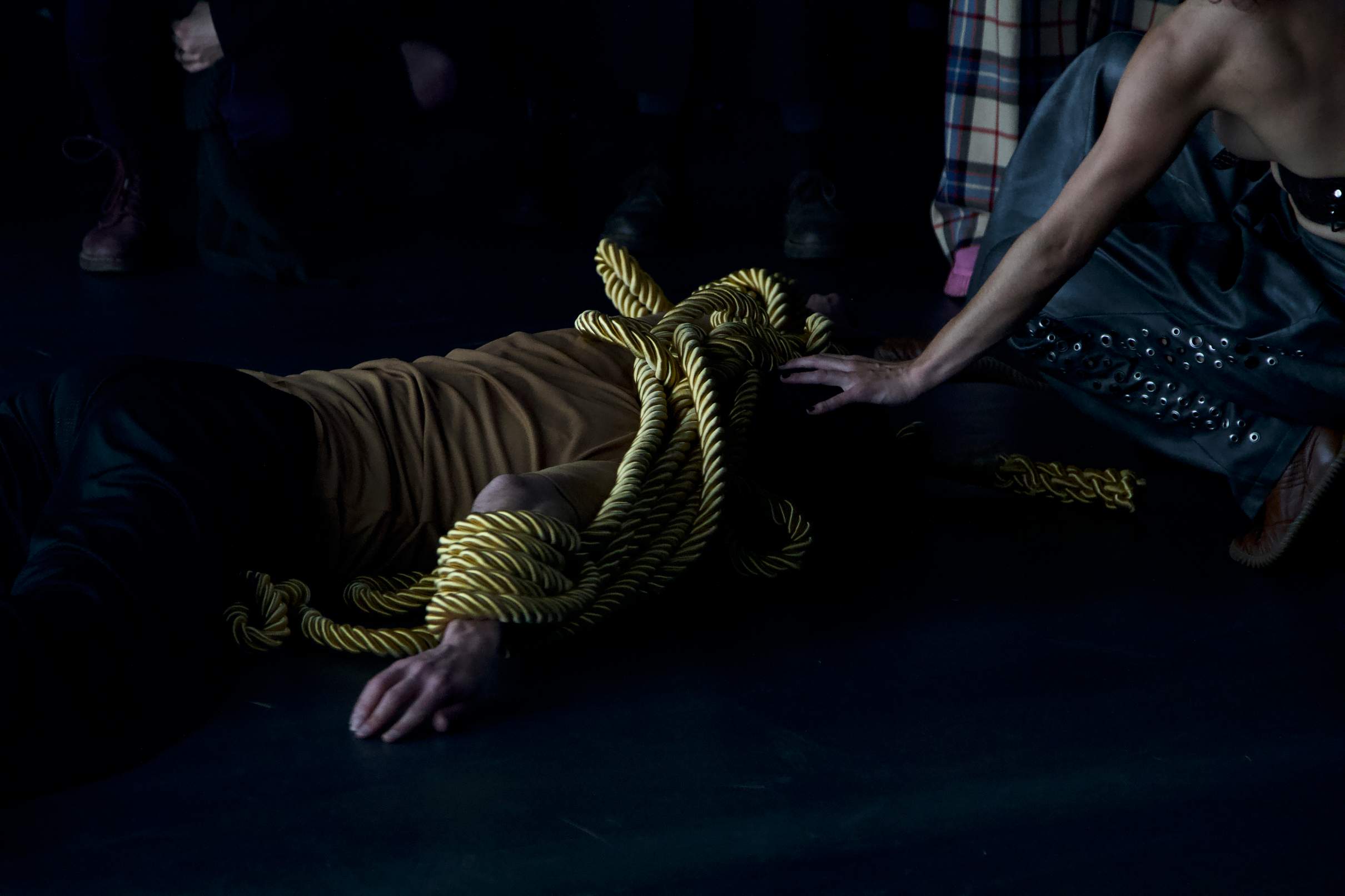 A performer lies prone on the floor, entangled in a thick golden rope around the wrist and shoulder.