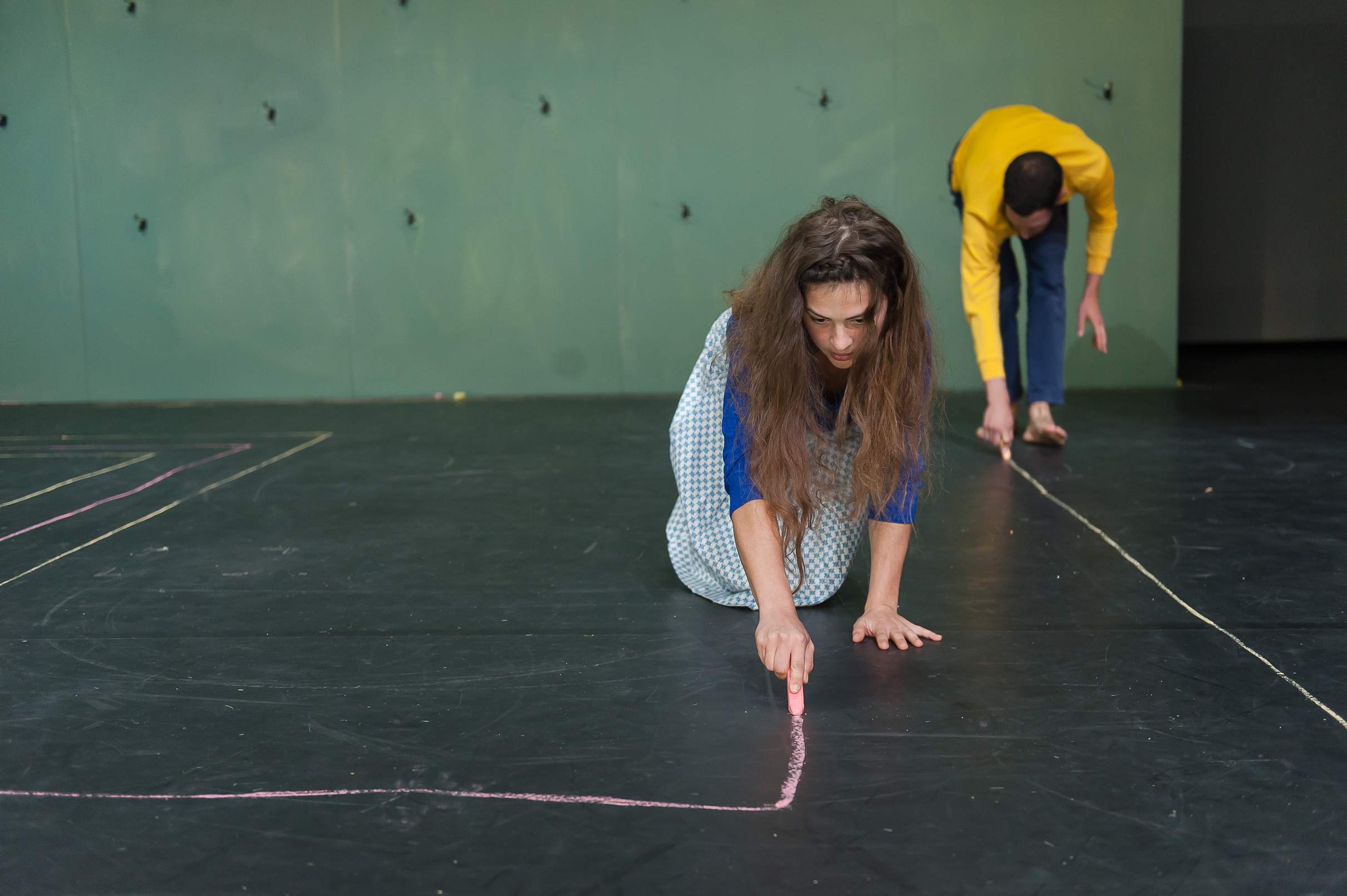 Two actors kneel or bend over as they draw a pattern on the stage floor with colourful chalk.