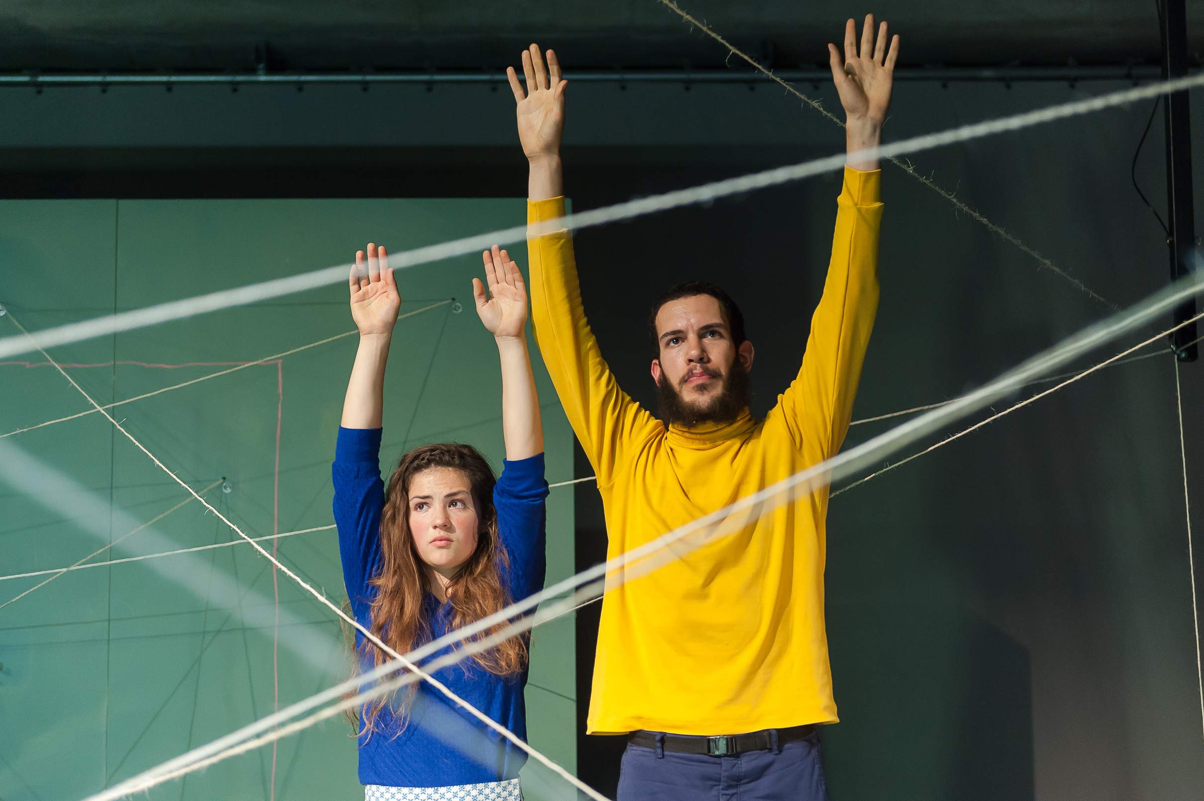 Two actors stand with hands raised between wires strung across the stage.