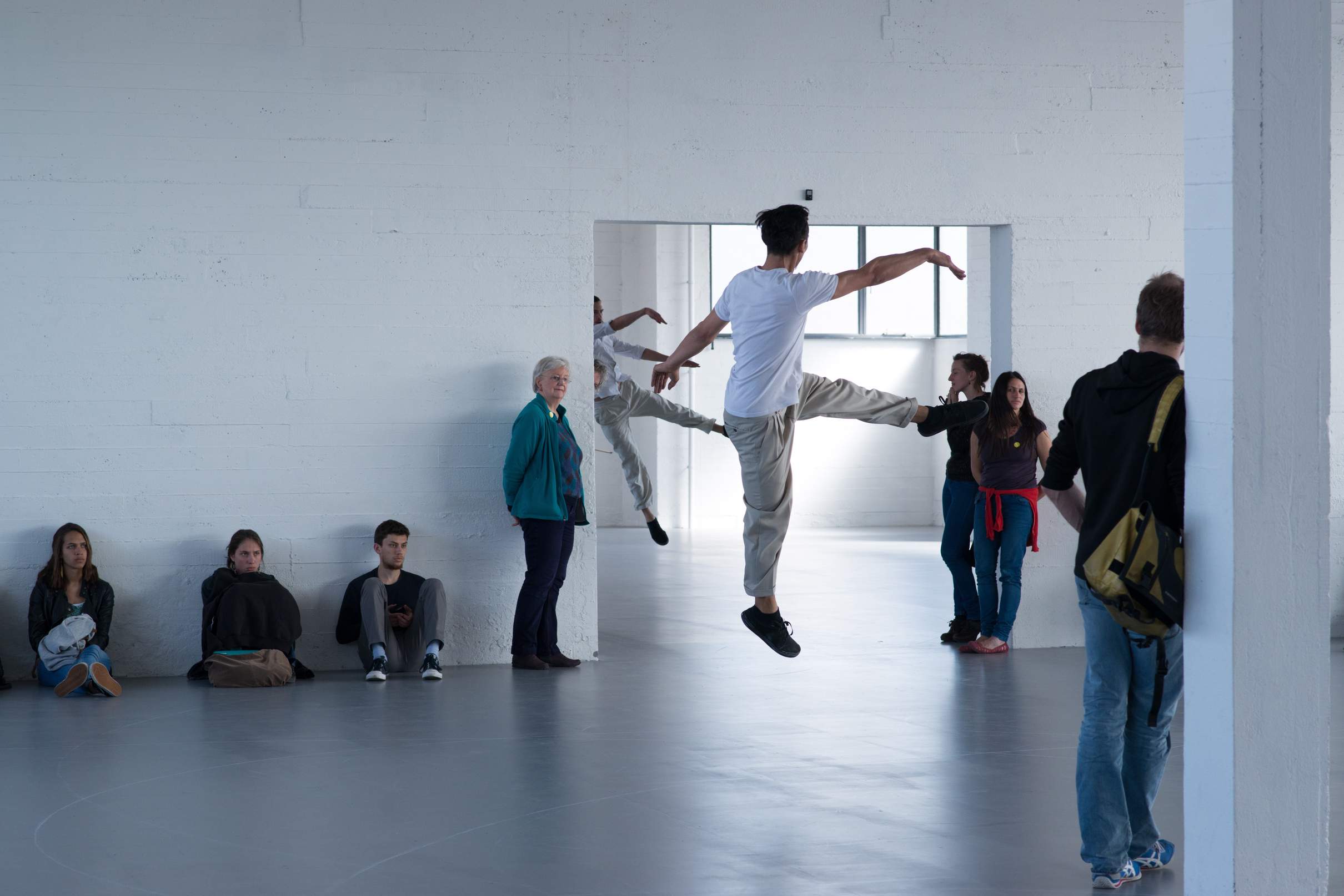 In two museum rooms connected by an opening, two dancers meet mid-jump in matching postures, looking at each other.